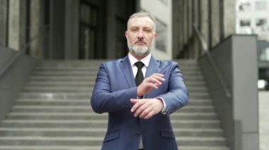 Portrait serious confident business mature man in formal suit looking at camera with crossed arms. Bearded gray-haired Businessman near office building outside stands on the urban street of the city