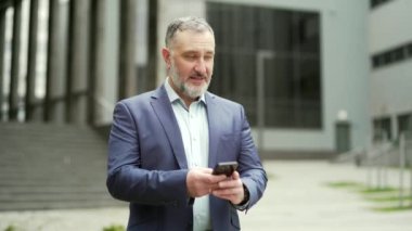 Successful senior investor with mobile phone outside office building Middle-aged businessman in business suit using smartphone, entrepreneur Chatting browsing, writing text message Outdoor city street