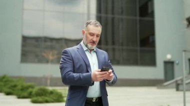 Confident business mature experienced gray-haired man in a formal suit walking down the city street with a smartphone phone in his hands against an urban background of an office building. outside