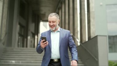 Confident business mature experienced gray-haired man in a formal suit walking down the city street with a smartphone phone in his hands against an urban background of an office building. outside