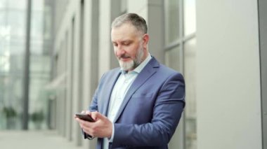 Happy mature business man in formal suit using smartphone while standing outside near the office building. Senior male employee browsing chatting uses mobile phone reading good news smiling. outdoor