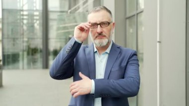 Portrait serious confident business mature man in formal suit looking at camera with crossed arms. Bearded gray-haired Businessman near office building outside stands on the urban street of the city