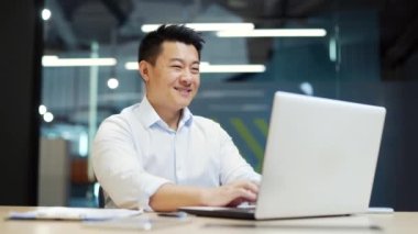 Asyalı ofis çalışanı işyerinde otururken kamerayı işaret ediyor. Beyaz gömlekli yakışıklı bir iş adamının portresi dizüstü bilgisayarda çalışıyor kameraya gülümsüyor