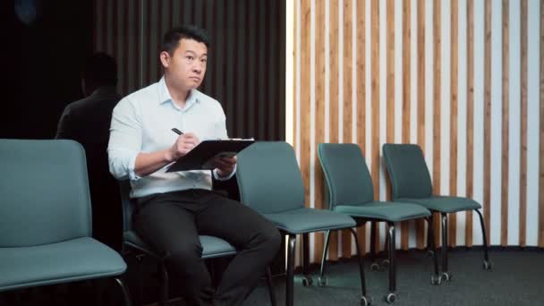 就職面接や会議のための待合室で若いアジア系男性 椅子に座っている間にフォルダ紙を持っている従業員の男はオフィスで履歴書の質問票を記入 — ストック動画
