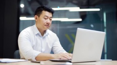 Sıkıcı Asyalı ofis çalışanı, iş yerinde bilgisayar masasında oturup elini uzatıyor. Yorgun adam. Yorgun bir iş adamı. Depresyonda, mutsuz, işsiz erkek menajer portresi. İç mekan