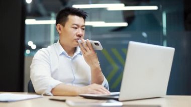 Modern ofiste çalışan ciddi bir Asyalı iş adamı dizüstü bilgisayarla yazı yazıyor, iş adamı sesli mesaj kaydediyor, telefondaki uygulamayla akıllı telefon hoparlöründen iletişim kuruyor. 