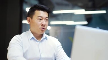 Asyalı erkek çalışanlar, ofiste dizüstü bilgisayarla otururken video aracılığıyla uzaktan iletişim kuruyor. İş adamı toplantısı, konferanslar, iş yerindeki iş arkadaşlarıyla görüşmeler.