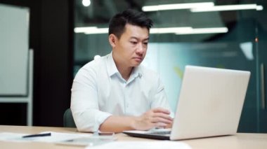 Düşünceli düşünceli Asyalı erkek çalışanın portresi, iş adamı yöneticisinin modern ofis masasındaki bilgisayarda uygulamalı tarama yapan bir finansal analist olduğunu düşünüyor.