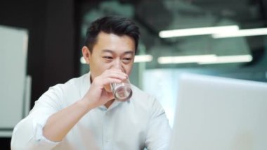 Asyalı çalışan, iş yerindeki modern ofiste çalışır dizüstü bilgisayarın başında oturup bardaktan temiz su içer. İş adamı, işçi, susuzluğunu giderir.
