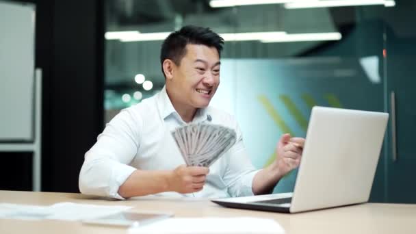 成功したアジアのビジネスマンがお金を持っている現金は画面のラップトップコンピュータを見て勝つ従業員を笑ってオフィスの机の上に座って笑顔で働く幸せなビジネスマンは収益を楽しむ — ストック動画