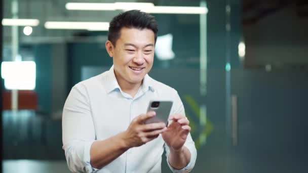 幸せなアジアの男性従業員は スマートフォンで良いニュースを読む成功を楽しんでいます ビジネスマンがモバイルオンライン賭けに勝つお金の入札財務賞の勝利は オフィスの屋内でスマートフォンで提供機会を受ける — ストック動画