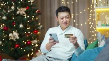 Asyalı adam yeni yıl ağacı arka planında cep telefonu ve kredi kartı kullanarak online alışveriş yapıyor. Kanepede oturup hediye almak telefon kullanarak internet siparişi vermek.