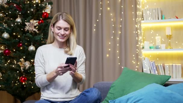 Γυναίκα Χρησιμοποιεί Κινητό Τηλέφωνο Κάθεται Στο Σπίτι Φόντο Χριστουγεννιάτικο Δέντρο — Αρχείο Βίντεο