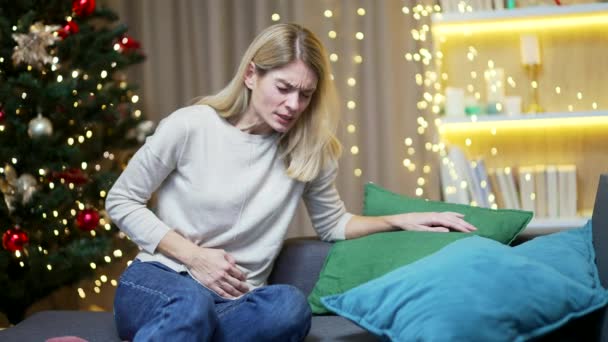 除夕夜 女人坐在沙发上 背靠着圣诞树 感到肚子痛和恶心 月经痛等感觉很糟 — 图库视频影像