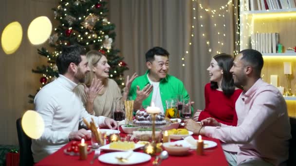 友人は一緒にテーブルに座ってクリスマスの前夜または新年の休日のお祝いを祝います 多民族のお祝いのクリスマスディナーのホームグループでの饗宴 幸せな明るい人は楽しそうに笑う — ストック動画