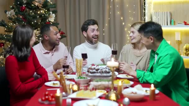 友人は一緒にテーブルに座ってクリスマスの前夜または新年の休日のお祝いを祝います 多民族のお祝いのクリスマスディナーのホームグループでの饗宴 幸せな陽気な人々クリンクワイングラスは笑う — ストック動画