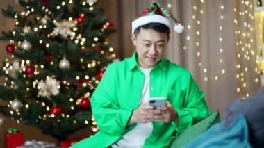 Neşeli Asyalı adam yeni yıl ağacı arka planında cep telefonuyla internetten alışveriş yapıyor. Daktilo, göz gezdirme, sohbet. Noel Baba şapkalı akıllı telefon erkeği. Gülümseme mesajı okur