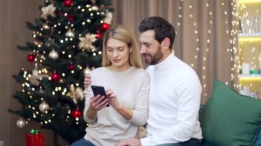 Mutlu evli çift, cep telefonuyla internet üzerinden yeni yıl alışverişi yapıyor. Akıllı telefon arka planıyla. Kanepede oturan bir Noel ağacı. Aile bakınıyor ve hediyeleri uzaktan seçiyor.