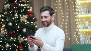 Yakışıklı, sakallı, beyaz bir adam Noel ağacının arka planında koltukta cep telefonu kullanıyor. Yazı yazan, sohbet eden, akıllı telefonlara bakan mutlu bir erkek. Çevrimiçi alımlar yapar, mesaj alışverişini okur
