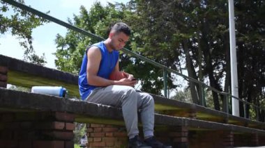 Mavi kolsuz tişörtlü mavi bir adam basketbol oynadıktan sonra dinleniyor ve telefonuna bakıyor.