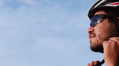 Bisiklet güneş gözlüğü takan sakallı bir adam ve arka planda mavi gökyüzü olan bisiklet kaskını emniyete alıyor. 