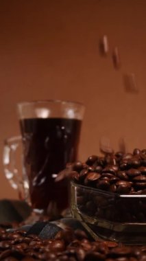 Bir fincan sade kahvenin yanında cam bir çaydanlıktan çıkan siyah kahve çekirdekleri.
