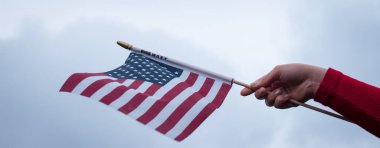 Kırmızı bir kazak giyiyor ve bulutlu bir arka planı olan bir ABD bayrağı sallıyor.