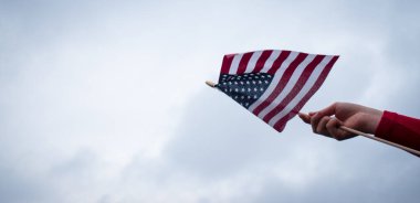 Kırmızı bir kazak giyiyor ve bulutlu bir arka planı olan bir ABD bayrağı sallıyor.