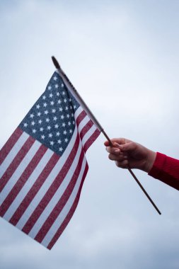 Kırmızı bir kazak giyiyor ve bulutlu bir arka planı olan bir ABD bayrağı sallıyor.