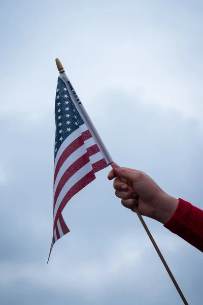 Kırmızı bir kazak giyiyor ve bulutlu bir arka planı olan bir ABD bayrağı sallıyor.