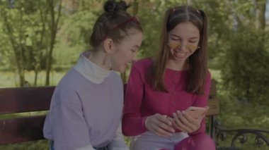 Kızlar parkta eğlenceli müzik dinleyerek ve bankta konuşarak vakit geçiriyorlar..
