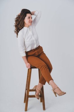 Genç bir kız beyaz arka planda bir sandalyeye oturur ve başka tarafa bakar. Saçlarını eliyle tutuyor..