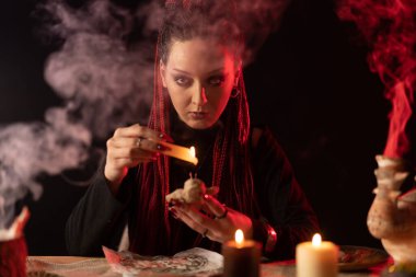 Cadı doğrudan kameraya bakar ve bir vudu bebeği tutar ve üzerine balmumu damlatır..