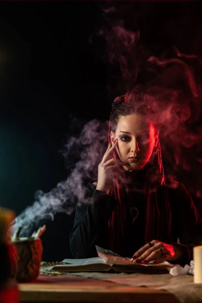 De heks zit aan tafel en houdt haar hand tegen haar gezicht. De rook van de kaars waait naar de heks.. — Stockfoto