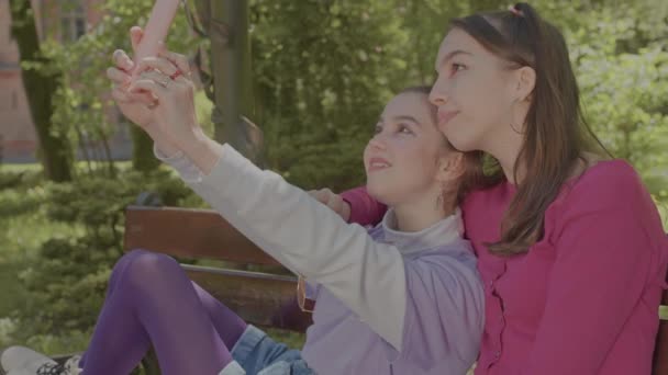Две младшие сестры делают селфи на скамейке в парке. Девушки улыбаются в камеру. — стоковое видео