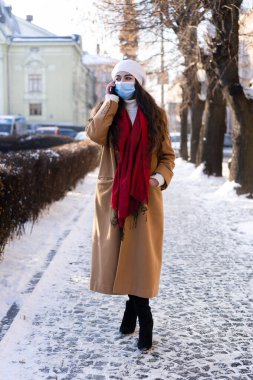 Genç ve güzel bir kızın fotoğrafı. Uzun kıvırcık saçlı, şık bir paltosu ve yüzünde maskesiyle. Karantinada yaşıyor.