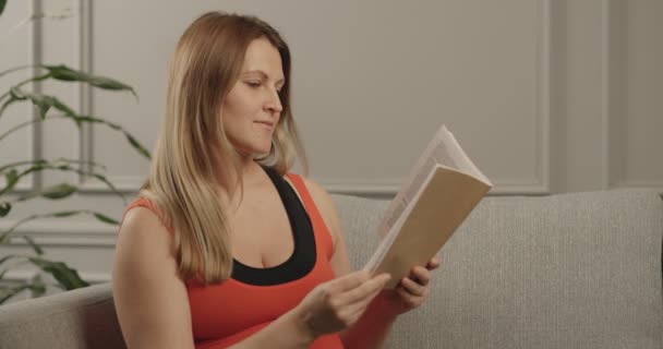 妊婦は出産に関する本を読んでいる。ソファに座り、赤いTシャツを着て. — ストック動画