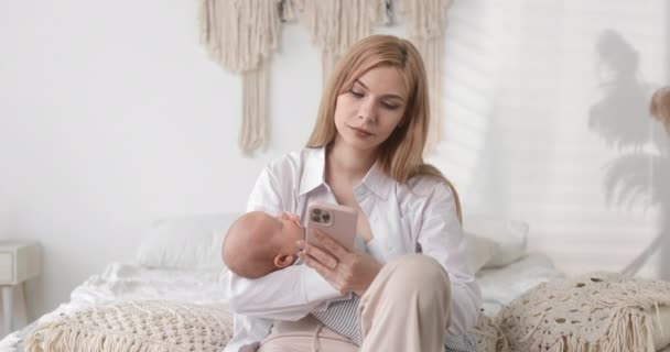 Mom holds the baby in her arms and works on the phone. A combination of work and motherhood. — Stock video