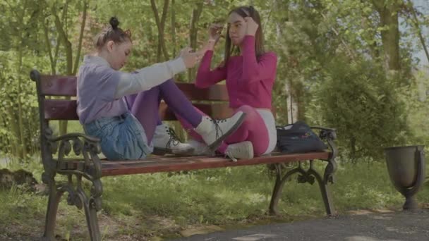 As meninas passam o tempo juntas ouvindo música divertida no parque e conversando no banco. — Vídeo de Stock