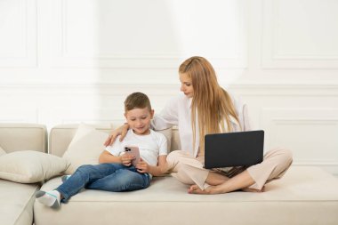 Anne ve oğlu kanepede telefonda oturuyor. Childs uzaktan öğreniyor ve anneler dizüstü bilgisayarda çalışıyor. Yatay fotoğraf.