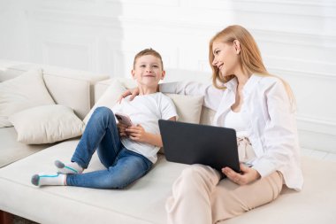 Oğlu telefonda annesine çeşitli videolar gösteriyor. Annem dizüstü bilgisayarda çalışıyor ve oğullarının videolarını izliyor. Odanın içi aydınlık..