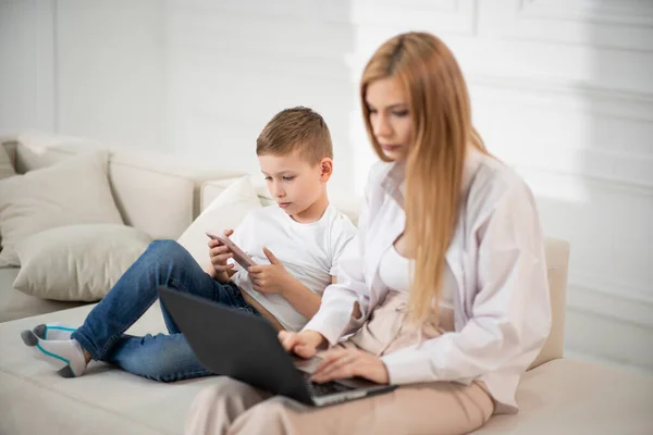 A busy mom works at a computer. Son shows funny videos to mom. Mom works and sits with the baby at home. Time in gadgets.