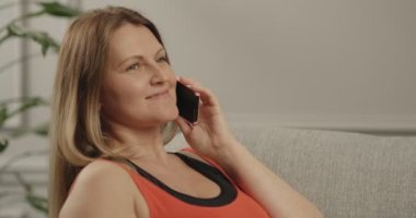 Telefonda konuşan ve mutlu bir şekilde gülümseyen bir kadın. O, odada oturuyor. Yatay video kapat.