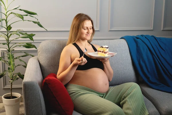 Kanepede oturup pasta yiyen mutlu hamile kadın. yatay resim.