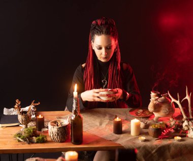 The witch sits at the table and looks into the bowl. Girl with red hair braided in pigtails and with red long nails. Horizontal photo.