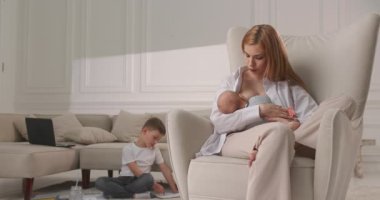 Anne, çocuğu sandalyeye oturtuyor ve en büyük oğul da annenin resmini çiziyor. Annem evde iki çocukla oturuyor..