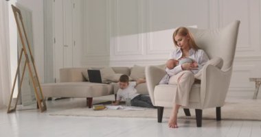 Annem bebeği sandalyede emziriyor. Arka planda, en büyük oğul resim çiziyor. Yatay video.