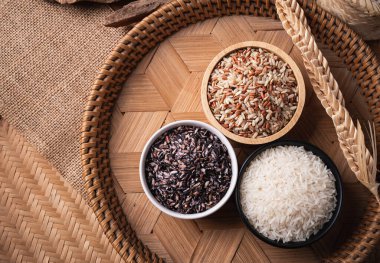 Bambu tepsisinde çeşitli organik Tayland pirinç taneleri (kahverengi, beyaz ve mor pirinç), masa üstü görünümü