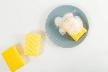 Gri tabakta sabunlu sarı bulaşık süngeri. Beyaz arka plan. Düz yat, boşluğu kopyala