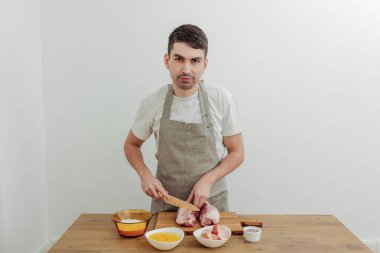 Önlüklü bir adam et pişiriyor. Yumurta, un, masaya tuz, kesme tahtası ve bıçak..